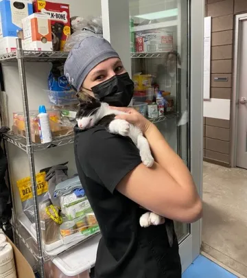 Ashley Sembach wears scrubs, a cap, and a mask and holds a black and white cat.