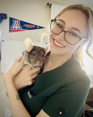A woman wearing scrubs holds a chinchilla and smiles.